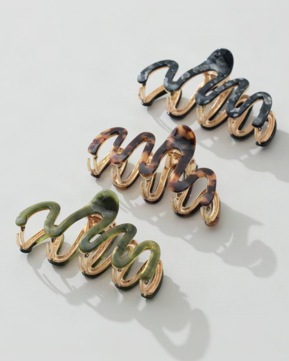 Three coiled hair claw clips arranged diagonally on a light gray background. The clips have glossy marbled enamel in black, tortoiseshell brown, and olive green over a gold-tone metal base.