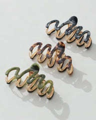 Three coiled hair claw clips arranged diagonally on a light gray background. The clips have glossy marbled enamel in black, tortoiseshell brown, and olive green over a gold-tone metal base.