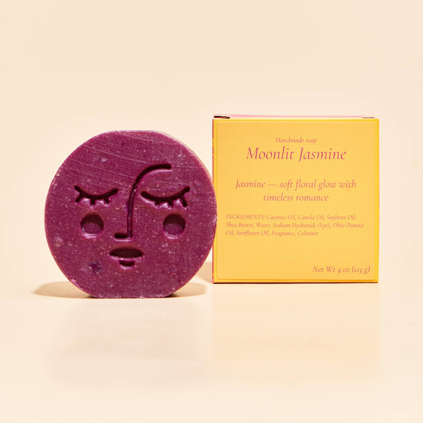 A round purple handmade soap bar with a moon face design sits next to its yellow packaging box labeled 'Moonlit Jasmine' on a warm cream background.