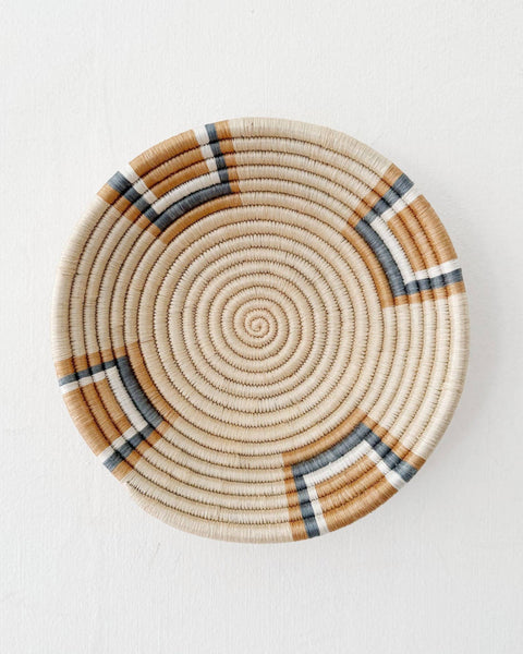 A round woven basket plate with a spiral pattern in natural beige, brown, and dark gray, photographed from directly above on a white background.