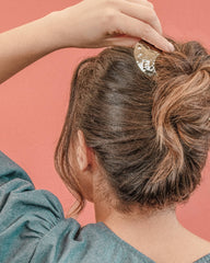 A close-up lifestyle shot of a woman from behind, demonstrating how to style her hair into a messy bun using a textured, metallic gold claw hair clip against a solid coral pink background.