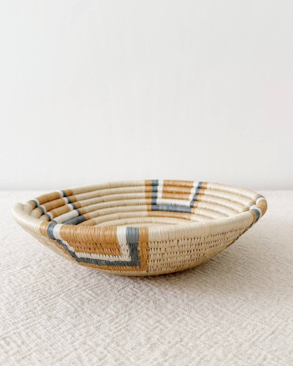 A shallow woven basket bowl with a natural beige base and geometric accents in white, brown, and gray, resting on a textured beige surface against a plain white wall.