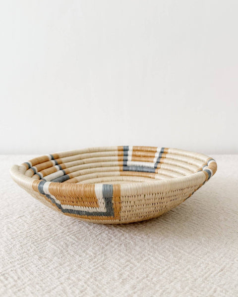A shallow woven basket bowl with a natural beige base and geometric accents in white, brown, and gray, resting on a textured beige surface against a plain white wall.