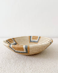 A shallow woven basket bowl with a natural beige base and geometric accents in white, brown, and gray, resting on a textured beige surface against a plain white wall.