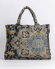 A structured tote bag featuring a distressed, vintage-style geometric and floral pattern in muted blue, gray, and ochre yellow, set against a plain light gray background.