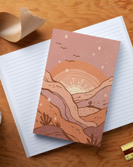 Notebook with a landscape design on a wooden surface with stationery items.