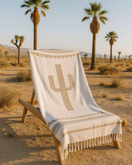 Beige blanket with cactus design draped over a wooden chair in a desert setting with palm trees.