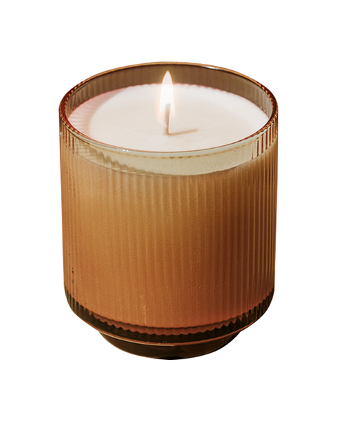 A lit candle in a ribbed amber glass jar with white wax, shown against a plain white background.