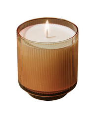 A lit candle in a ribbed amber glass jar with white wax, shown against a plain white background.