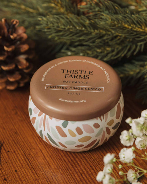 A Thistle Farms 'Frosted Gingerbread' soy candle in a white ceramic jar with a brown lid, styled on a wooden surface with pine branches, a pinecone, and baby's breath flowers.