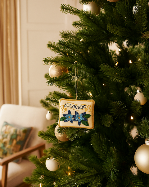 A square Christmas ornament hanging on a green Christmas tree. The ornament is cream-colored with the word 'COLORADO' in gold lettering and features blue and green floral designs.