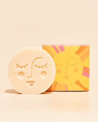 A round off-white soap bar with a sun face embossed on it, standing in front of its colorful sun-themed packaging box on a warm beige background.