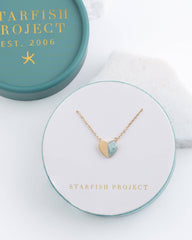 A gold heart pendant necklace with a light blue stone inlay, displayed on a white circular card with the Starfish Project logo, next to its teal presentation box.