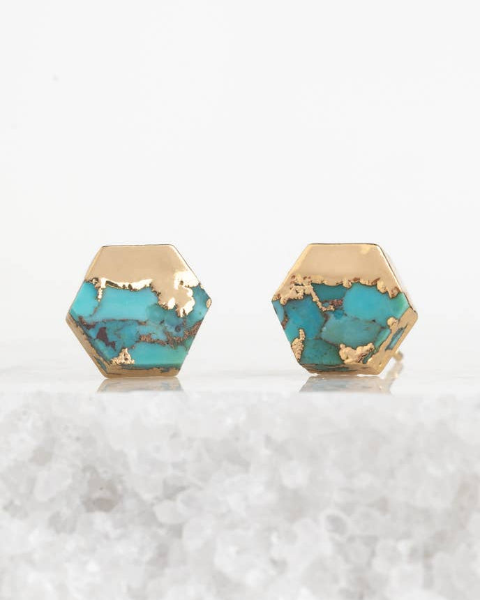 Hexagonal stud earrings with a gold-tone metal base and a turquoise blue stone inlay, displayed on a white textured surface against a white background.