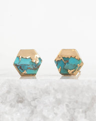 Hexagonal stud earrings with a gold-tone metal base and a turquoise blue stone inlay, displayed on a white textured surface against a white background.
