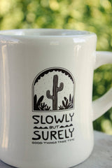 White mug with a cactus design and 'Slowly But Surely' text against a blurred green background