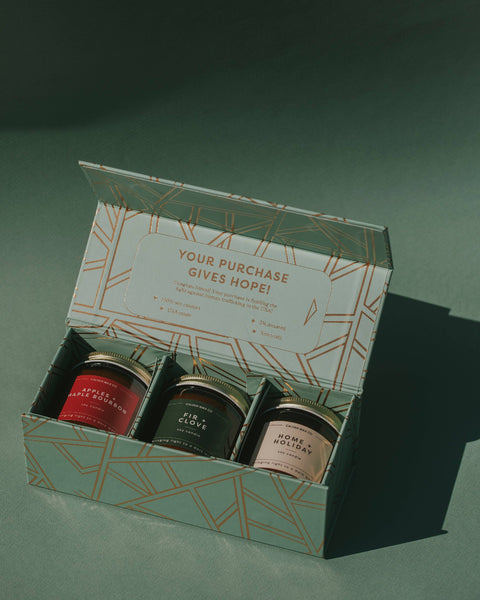 An open gift box containing three small jars with gold lids, set on a dark green surface with a soft shadow. The box lid has a geometric pattern and the text 'YOUR PURCHASE GIVES HOPE!'.
