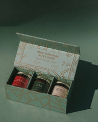 An open gift box containing three small jars with gold lids, set on a dark green surface with a soft shadow. The box lid has a geometric pattern and the text 'YOUR PURCHASE GIVES HOPE!'.