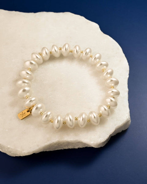 A pearl beaded bracelet with small gold spacer beads and a gold clasp, displayed on a white textured stone against a dark blue background.