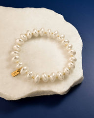 A pearl beaded bracelet with small gold spacer beads and a gold clasp, displayed on a white textured stone against a dark blue background.