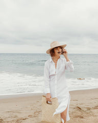 A woman stands on a sandy beach wearing a long white button-up shirt dress, a beige wide-brim hat, and holding a large woven straw tote bag. The ocean and cloudy sky are visible in the background.