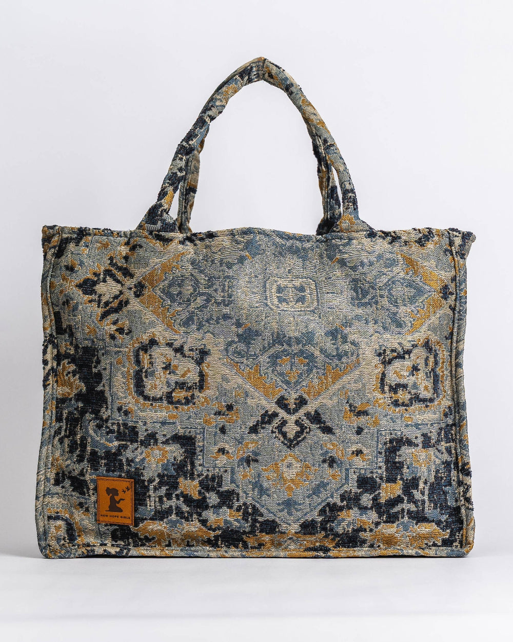 A rectangular tote bag with a distressed, vintage-style geometric pattern in navy blue, gray, and ochre yellow. It features two top handles and a small brown leather brand patch on the lower left front.