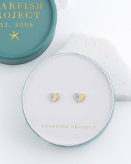 Two small heart-shaped stud earrings with a gold and light blue color combination, displayed on a round white jewelry stand with the brand name 'STARFISH PROJECT' printed in gold. The stand is placed on a white surface next to its teal and gold branded box.