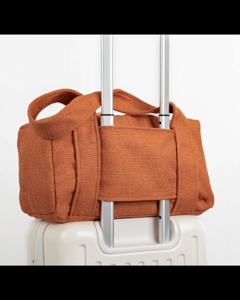 An orange-brown canvas duffel bag with top handles, shown strapped around the handle of a white rolling suitcase against a plain white background.