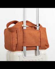 An orange-brown canvas duffel bag with top handles, shown strapped around the handle of a white rolling suitcase against a plain white background.