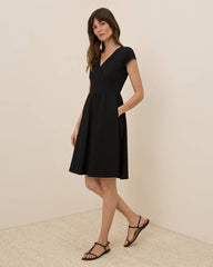 A woman modeling a black short-sleeve wrap-style dress with a flared skirt and side pockets, paired with black strappy sandals, against a warm beige studio background.