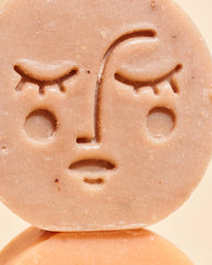 A close-up of a round, light brown handmade soap bar featuring an embossed face design, resting on a matching surface against a warm beige background.