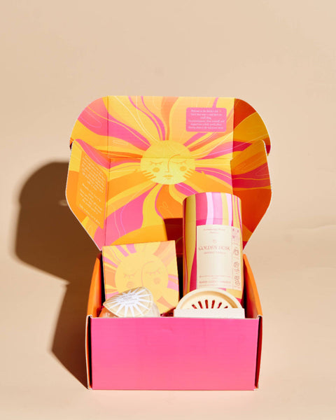 An open gift box with a bright pink base and a sunburst-patterned lid in orange, yellow, and pink, containing several small toiletry or bath products on a warm beige background.