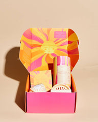 An open gift box with a bright pink base and a sunburst-patterned lid in orange, yellow, and pink, containing several small toiletry or bath products on a warm beige background.