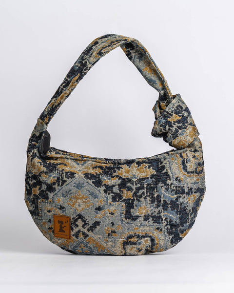 A hobo-style shoulder bag with a distressed, vintage-inspired geometric and floral pattern in navy blue, beige, and muted gold, featuring a small brown leather brand patch on the front.