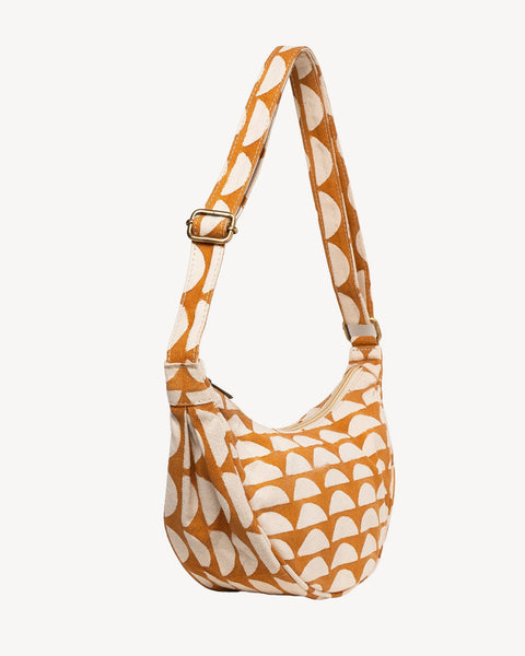 A slouchy shoulder bag with a warm orange and cream semi-circular pattern, featuring an adjustable shoulder strap with a gold-tone buckle, set against a solid off-white background.