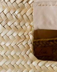 Close-up of a woven straw or raffia bag corner showing the natural beige texture, a stitched leather trim, and part of the interior.