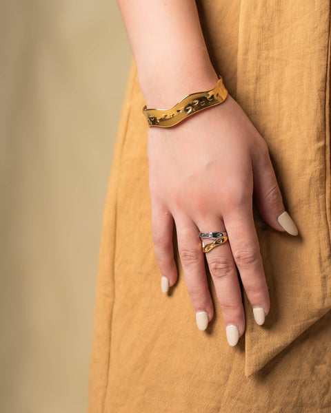 A close-up of a person's hand resting on a mustard yellow linen dress, wearing a gold chain-link bracelet on the wrist and a gold ring with a blue stone on the finger.