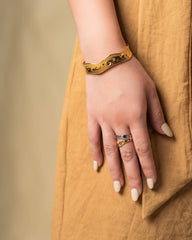 A close-up of a person's hand resting on a mustard yellow linen dress, wearing a gold chain-link bracelet on the wrist and a gold ring with a blue stone on the finger.