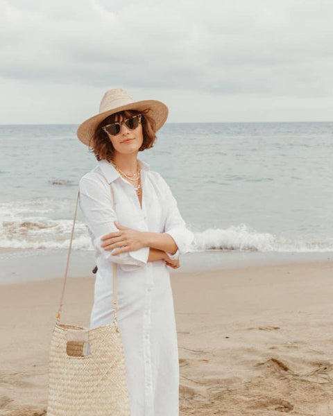 A woman stands on a sandy beach wearing a wide-brim straw hat, white long-sleeve button-up shirt dress, sunglasses, layered necklaces, and carrying a large woven straw tote bag.