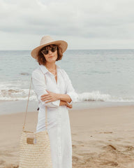 A woman stands on a sandy beach wearing a wide-brim straw hat, white long-sleeve button-up shirt dress, sunglasses, layered necklaces, and carrying a large woven straw tote bag.