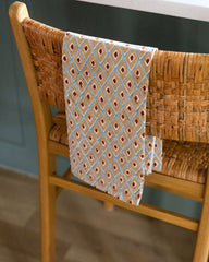 A patterned kitchen towel with a geometric diamond design in light blue, rust orange, and cream, draped over the back of a wooden woven chair.
