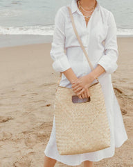 A woman standing on a sandy beach wearing a white long-sleeve shirt and white midi dress, carrying a large woven straw tote bag with a beige shoulder strap.