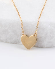 A gold heart-shaped pendant necklace engraved with the word 'hope', resting on a white textured stone surface.