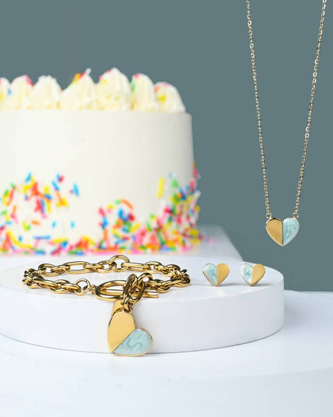 A gold heart-themed jewelry set displayed on a white pedestal, featuring a gold chain necklace with a split gold and light blue heart pendant, a gold link bracelet with matching heart charms, and small heart earrings. A white frosted cake with colorful sprinkles sits in the softly blurred background against a muted blue-gray wall.