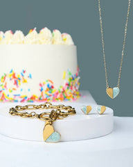 A gold heart-themed jewelry set displayed on a white pedestal, featuring a gold chain necklace with a split gold and light blue heart pendant, a gold link bracelet with matching heart charms, and small heart earrings. A white frosted cake with colorful sprinkles sits in the softly blurred background against a muted blue-gray wall.