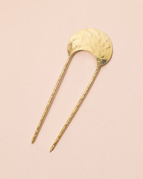 A gold-toned metal hair pin with a hammered circular top and two long twisted stems, photographed on a soft pink background.