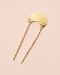 A gold-toned metal hair pin with a hammered circular top and two long twisted stems, photographed on a soft pink background.