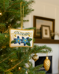 A decorative Christmas ornament shaped like a small sign reading "COLORADO" hangs on a green Christmas tree. The sign has a cream background with blue and green floral motifs and gold lettering, while the tree is lit with warm white lights and has gold pinecone ornaments nearby.