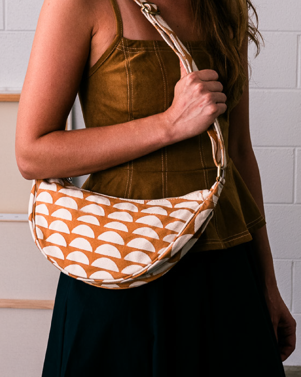 A woman wearing a brown camisole and black skirt carries a crescent-shaped shoulder bag with a terracotta orange and cream semicircle pattern against a white brick wall.