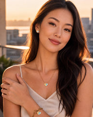 A woman with long dark hair wearing a gold heart pendant necklace, matching bracelet, and stud earring, posing outdoors during sunset with a blurred city skyline in the background.